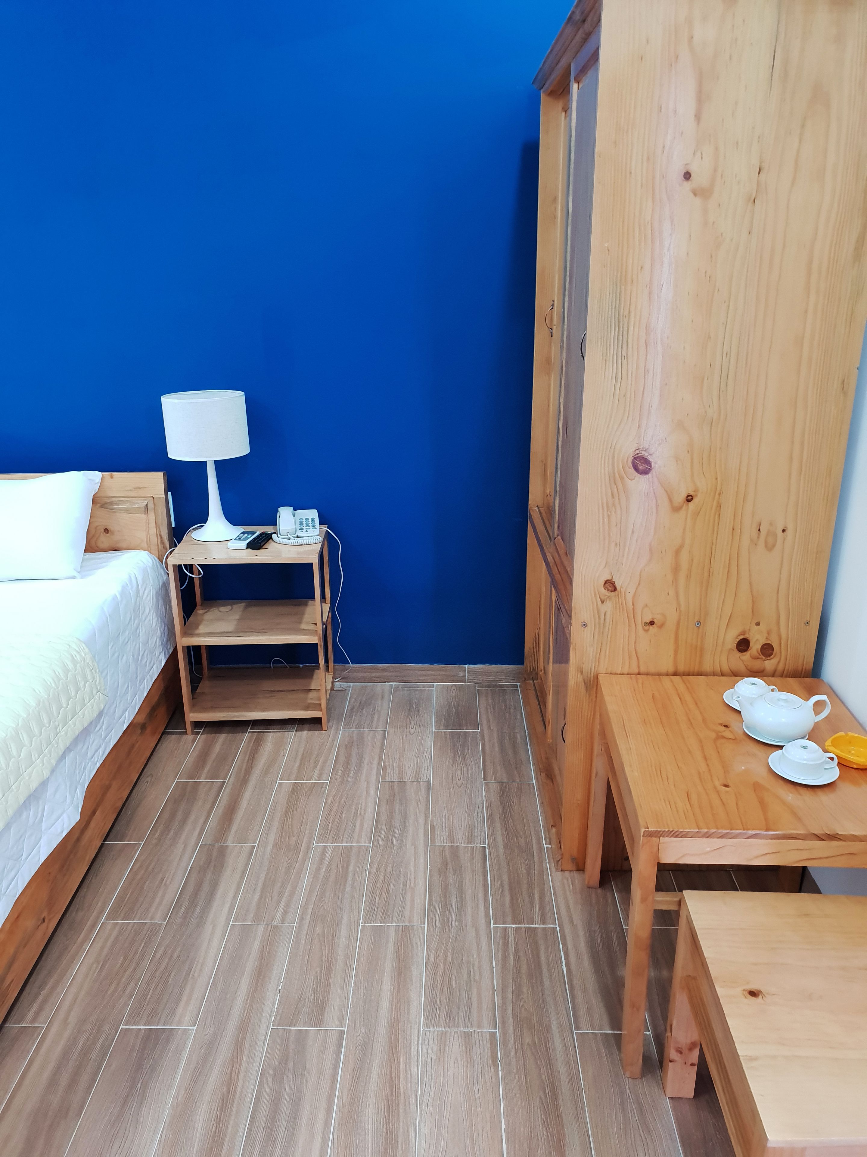 A bedroom with a bed, bedside table with a lamp, a small wooden shelf, and a wooden cabinet with tea set.