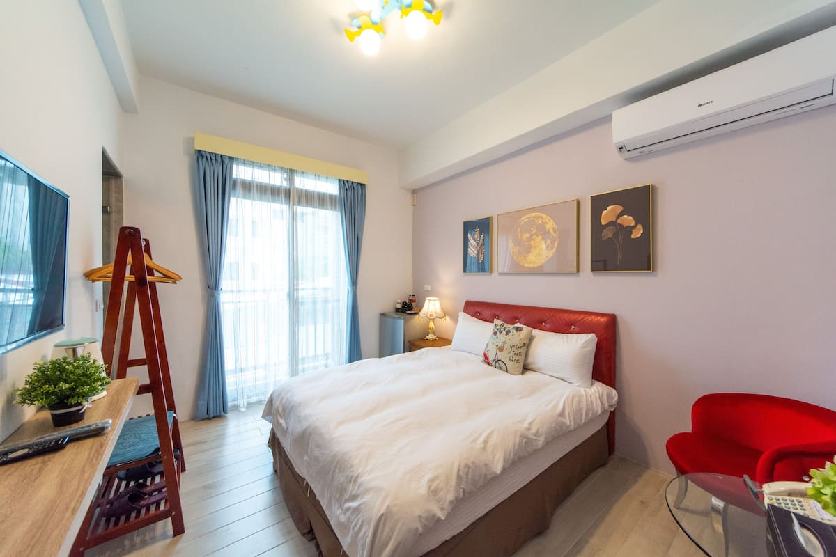 A modern hotel room with a large bed, a red chair, a desk with a lamp, and artwork on the walls.