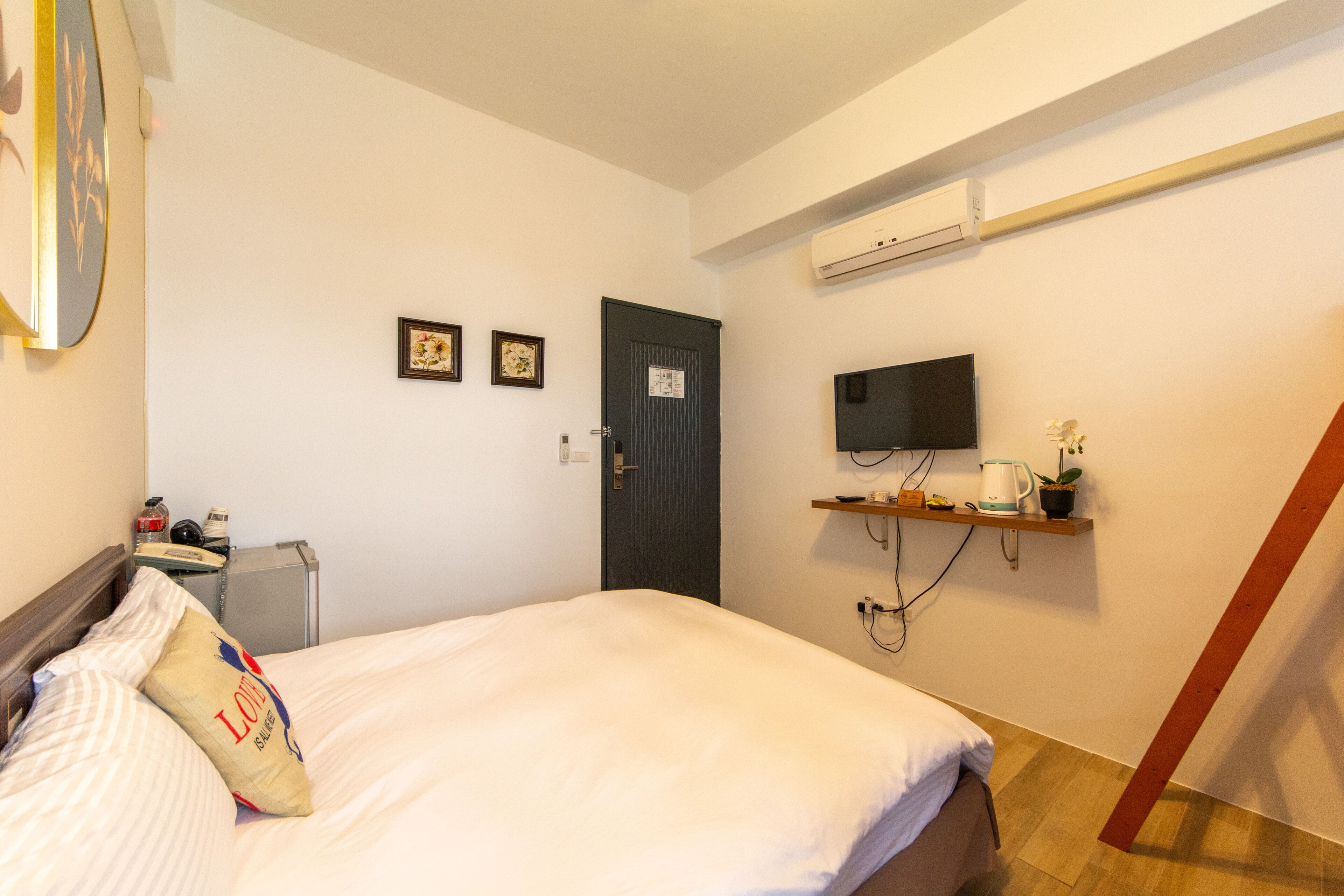 Habitación de hotel con una cama, un televisor montado en la pared, un pequeño estante con una planta y una tetera, y una puerta con un cartel.