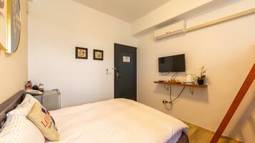 Habitación de hotel con una cama, un televisor montado en la pared, un pequeño estante con una planta y una tetera, y una puerta con un cartel.