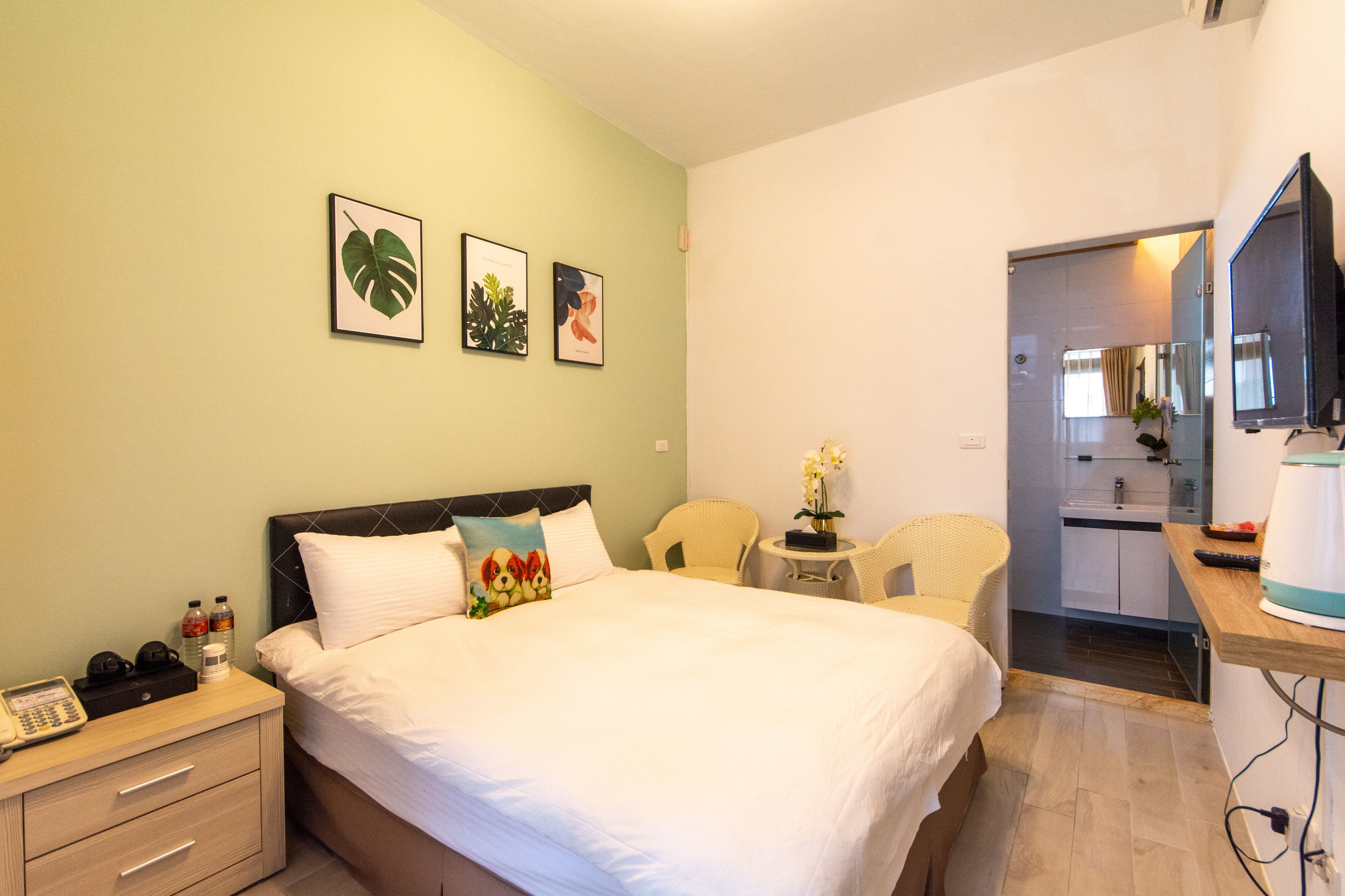 A hotel room with a bed, bedside table, and a small desk with a chair. There are framed pictures on the wall and a kitchen area visible through an open door.
