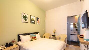 A hotel room with a bed, bedside table, and a small desk with a chair. There are framed pictures on the wall and a kitchen area visible through an open door.