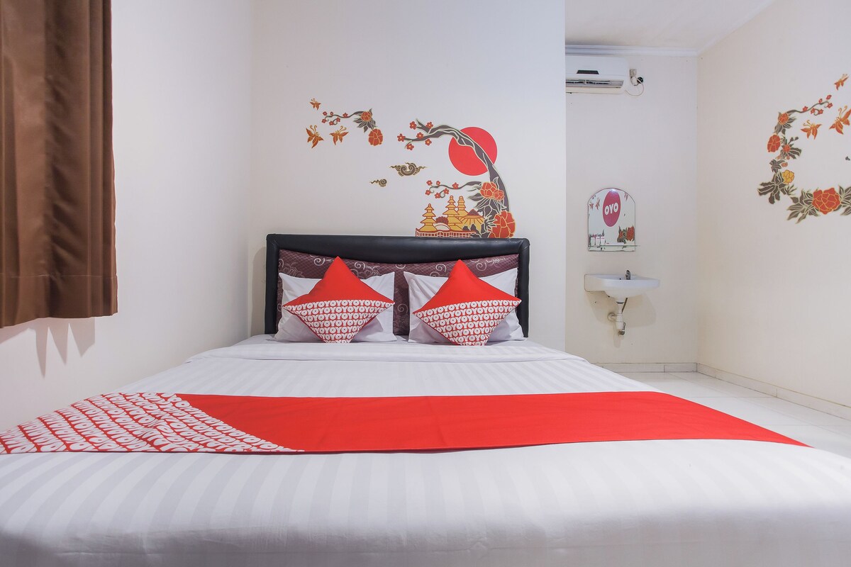 A hotel room with a bed, red and white pillows, a red throw blanket, a wall-mounted sink, and decorative wall stickers.