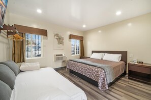 Ein Hotelzimmer mit einem großen Bett, einem Einzelbett, einem Schreibtisch und einem Sessel. Zwei Fenster mit Vorhängen, eine Wandklimaanlage und ein gerahmtes Bild sind ebenfalls vorhanden.