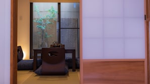 Ett traditionellt japanskt rum med tatamimattor på golvet, ett lågt bord med en mörk trästol och en skjutdörr med utsikt över bambu utanför.