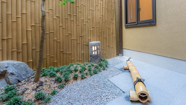 En traditionell japansk trädgård med ett bambustaket, ett litet träd, en stenlykta och en trätrumma.