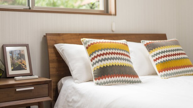 Un lit bien fait, avec du linge de lit blanc et des coussins aux motifs colorés en zigzag. Une petite table de chevet en bois, sur laquelle se trouvent un cadre avec une photo et une télécommande, est placée à côté du lit.
