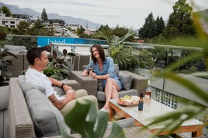 A couple seated on a wicker sofa with a table of snacks and drinks in a terrace setting with a city view.