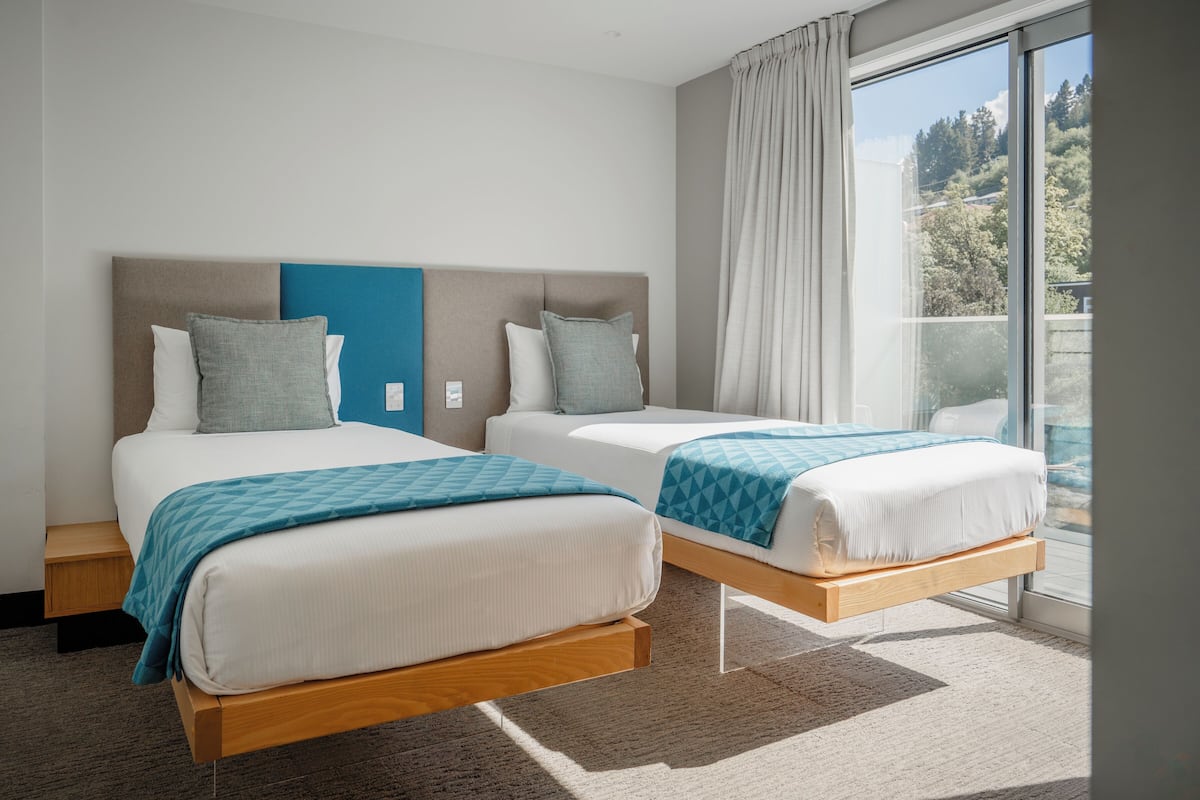 Quarto de hotel com duas camas, uma janela grande com cortinas e vista para árvores.