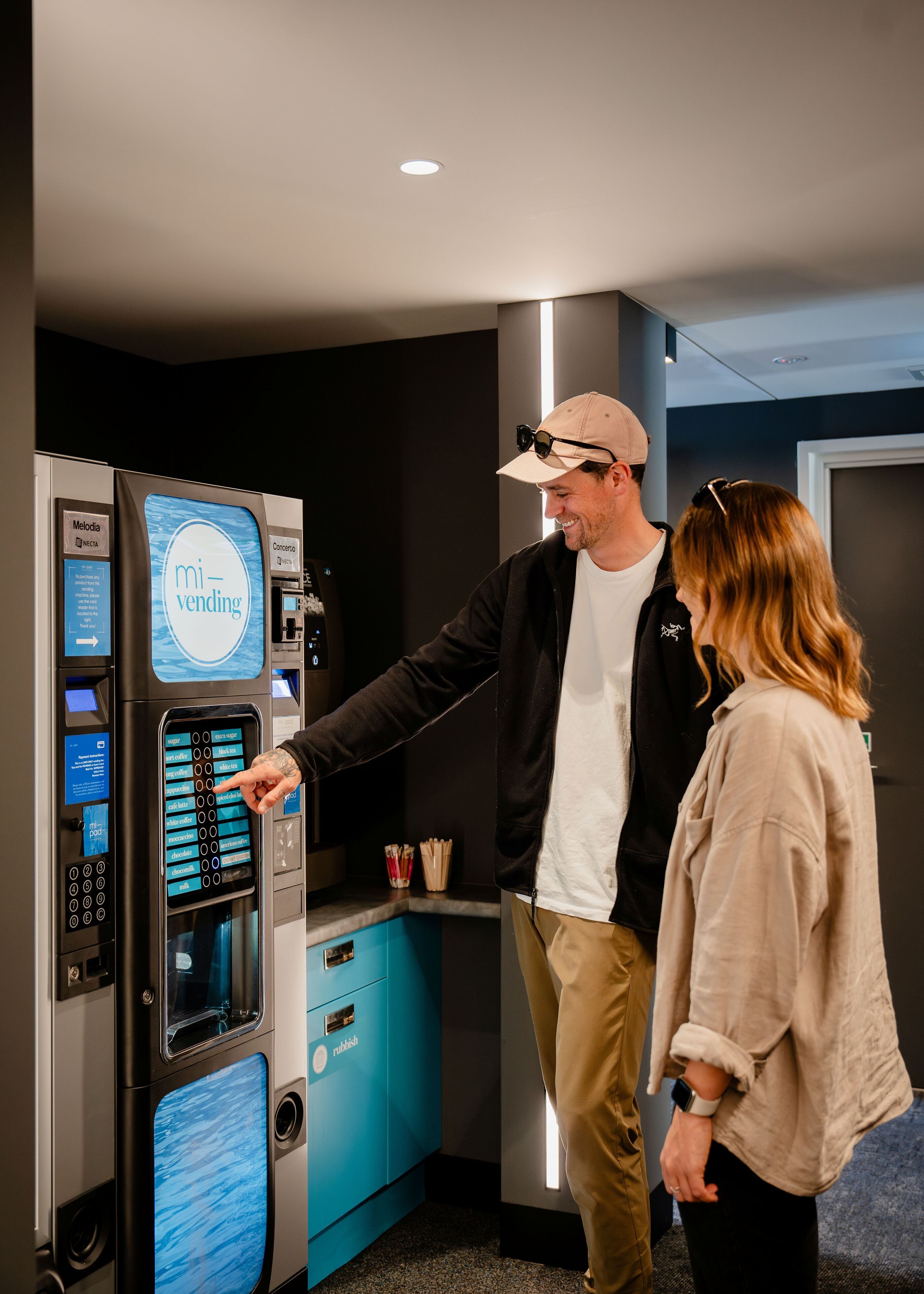 Una máquina expendedora con el rótulo "mi-vending", una pantalla táctil, una cafetera y un mueble azul con la etiqueta "rubish".