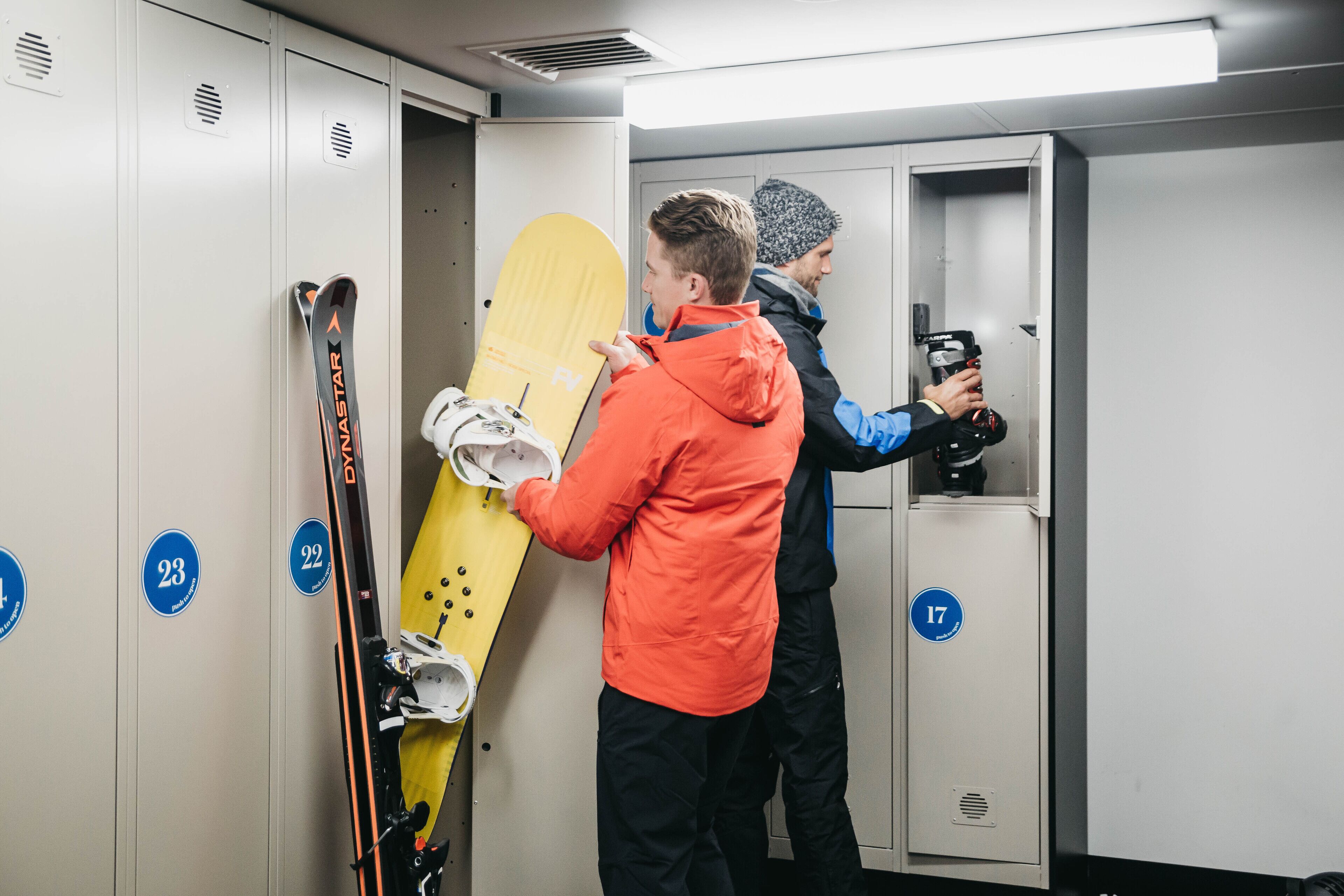 A locker room with numbered lockers, a yellow snowboard, and a person holding skis.