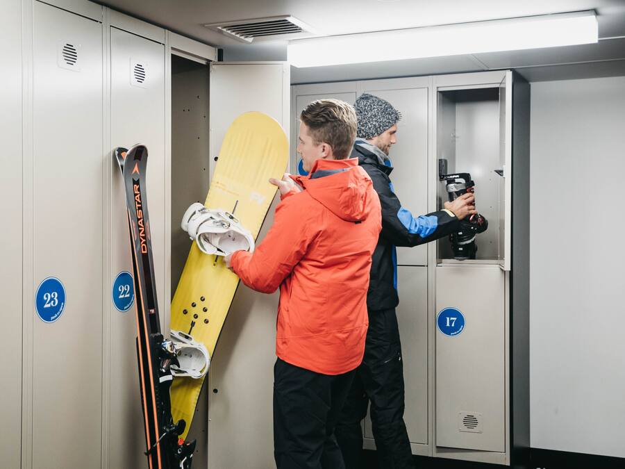 A locker room with numbered lockers, a yellow snowboard, and a person holding skis.