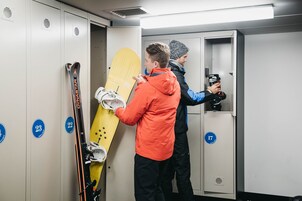 Vestiário com armários numerados, uma prancha de snowboard amarela e uma pessoa segurando esquis.