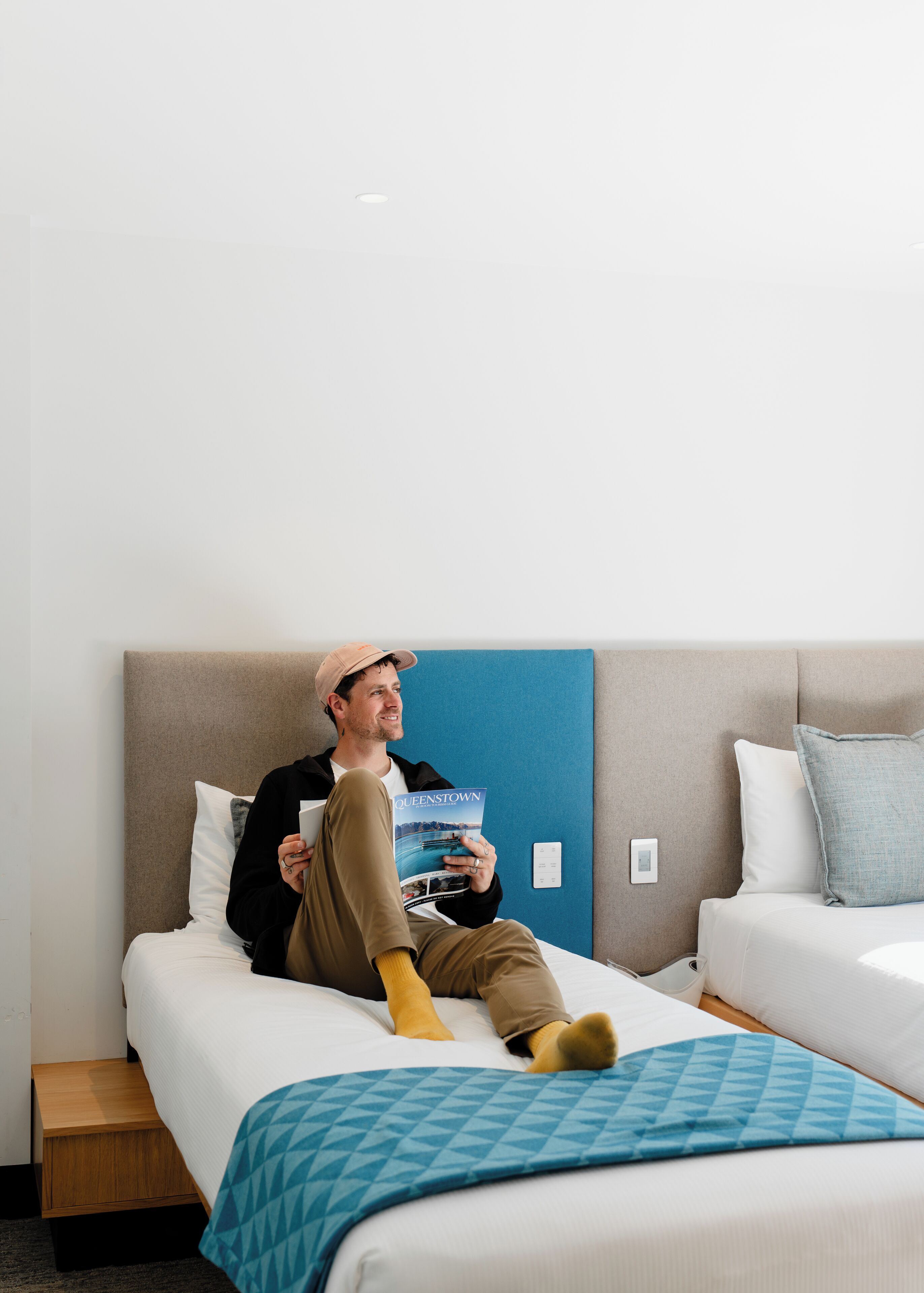 Uma pessoa sentada em uma cama em quarto de hotel com parede azul, lendo um livro intitulado 'Queenstown'.