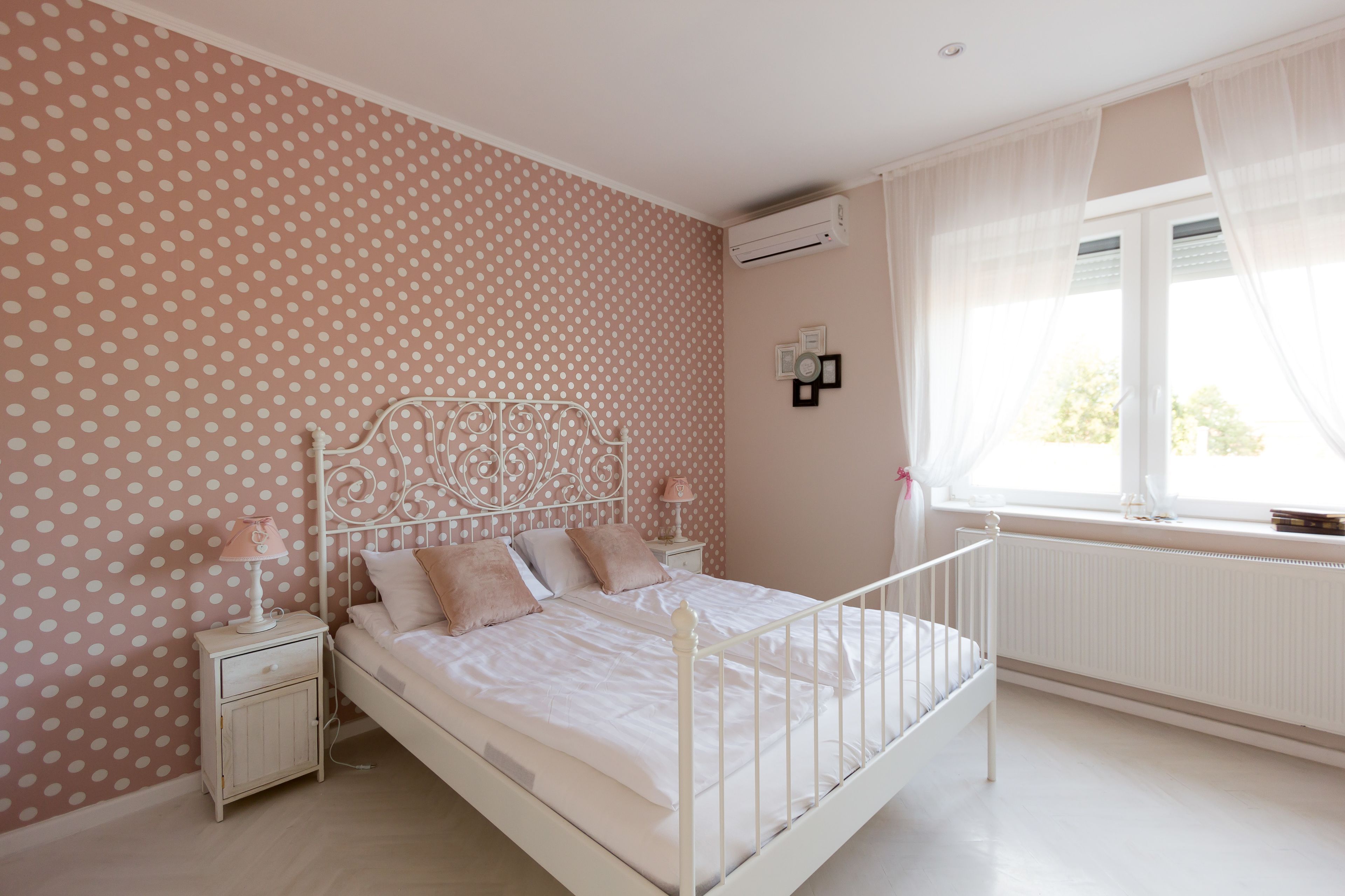 A bedroom with a polka dot wallpaper, a white bed with a metal headboard, a bedside table with a lamp, and a window with sheer curtains.