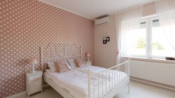 A bedroom with a polka dot wallpaper, a white bed with a metal headboard, a bedside table with a lamp, and a window with sheer curtains.