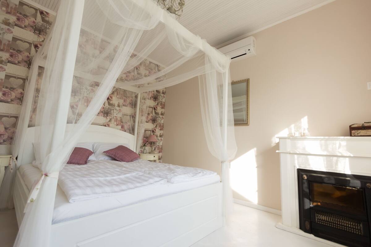 A bedroom with a four-poster bed, floral wallpaper, a white fireplace, and a mirror on the wall.