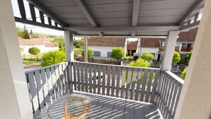 A covered balcony with a glass table and wicker chairs, overlooking a residential area with houses and trees.