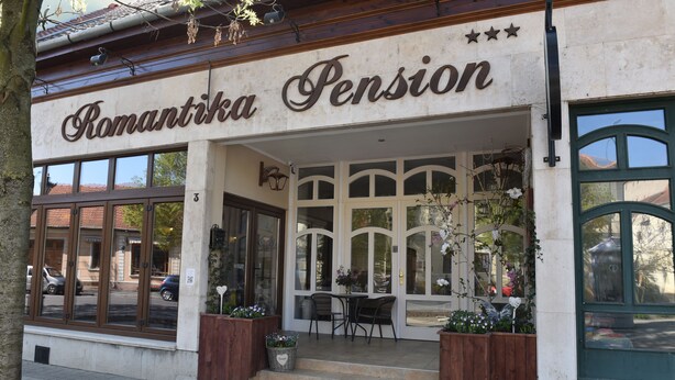 A building with a sign reading "Romantika Pension." It features large windows, a small outdoor seating area with tables and chairs, and potted plants.