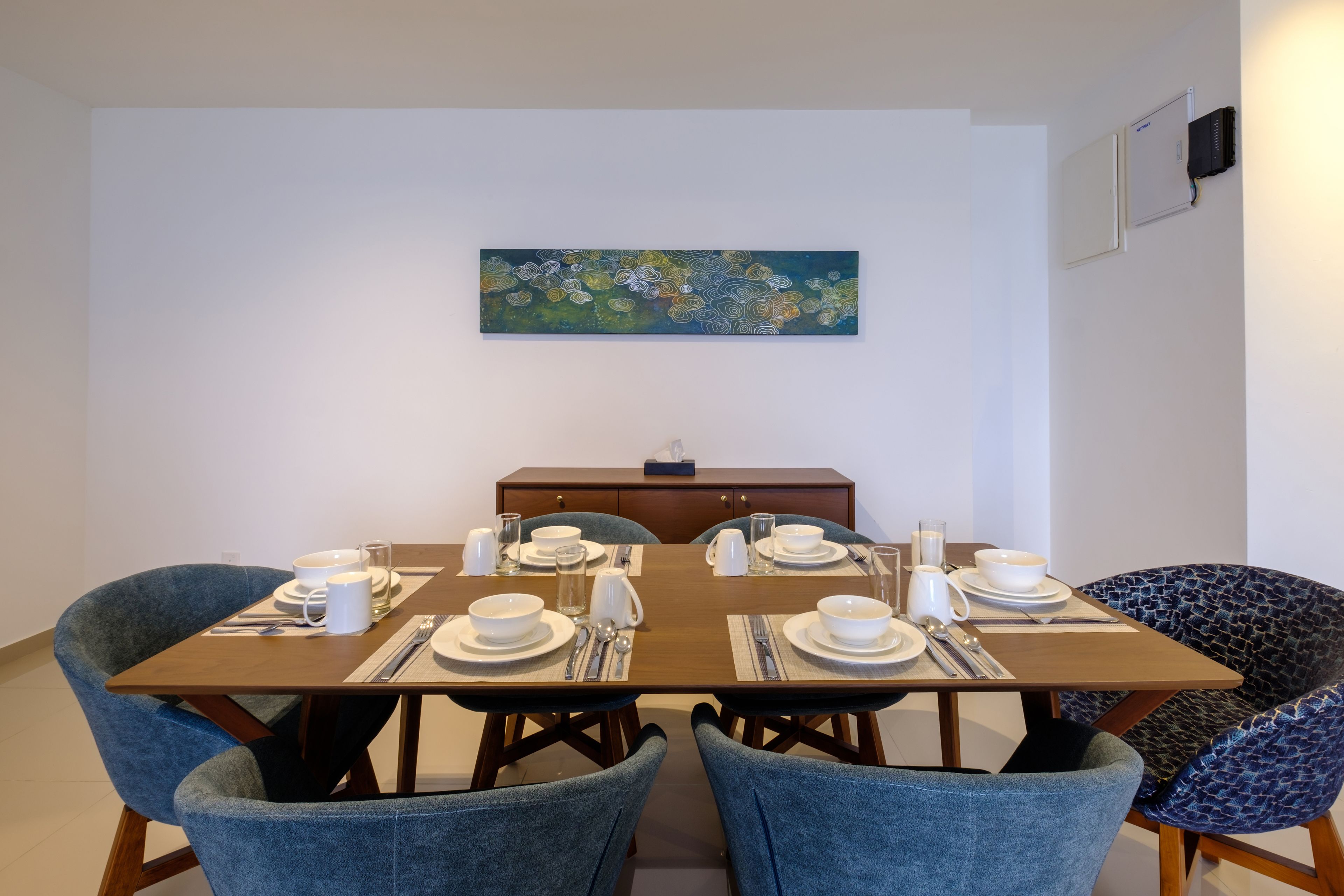 Uma sala de jantar com uma mesa retangular posta para quatro pessoas, cercada por quatro cadeiras azuis. Um quadro está pendurado na parede acima da mesa.