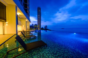 An infinity pool with lounge chairs, a modern building with balconies, and a view of the sea and a tall building in the background.