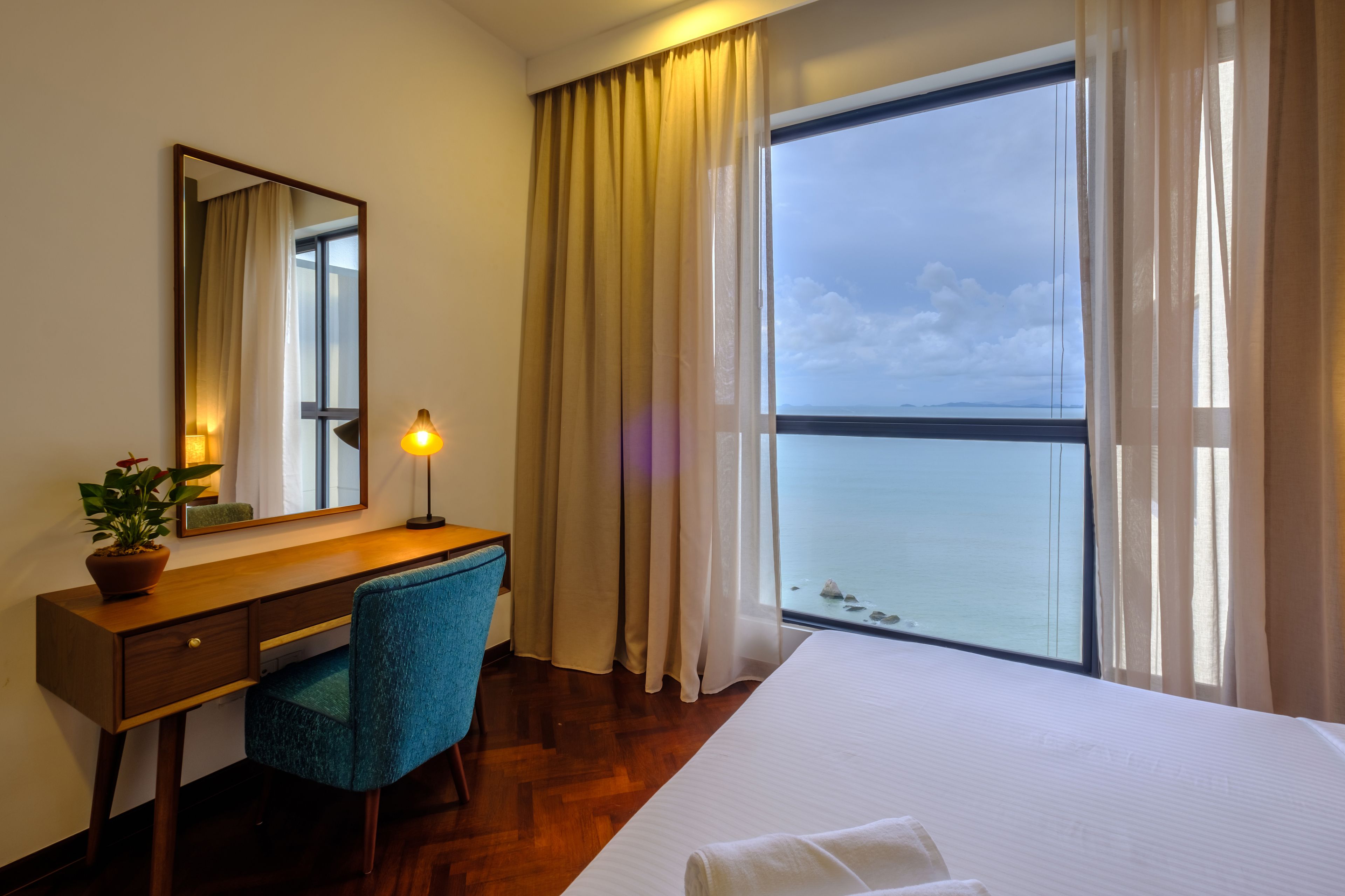 A hotel room with a large window, a bed, a desk with a chair, a mirror, and a lamp.
