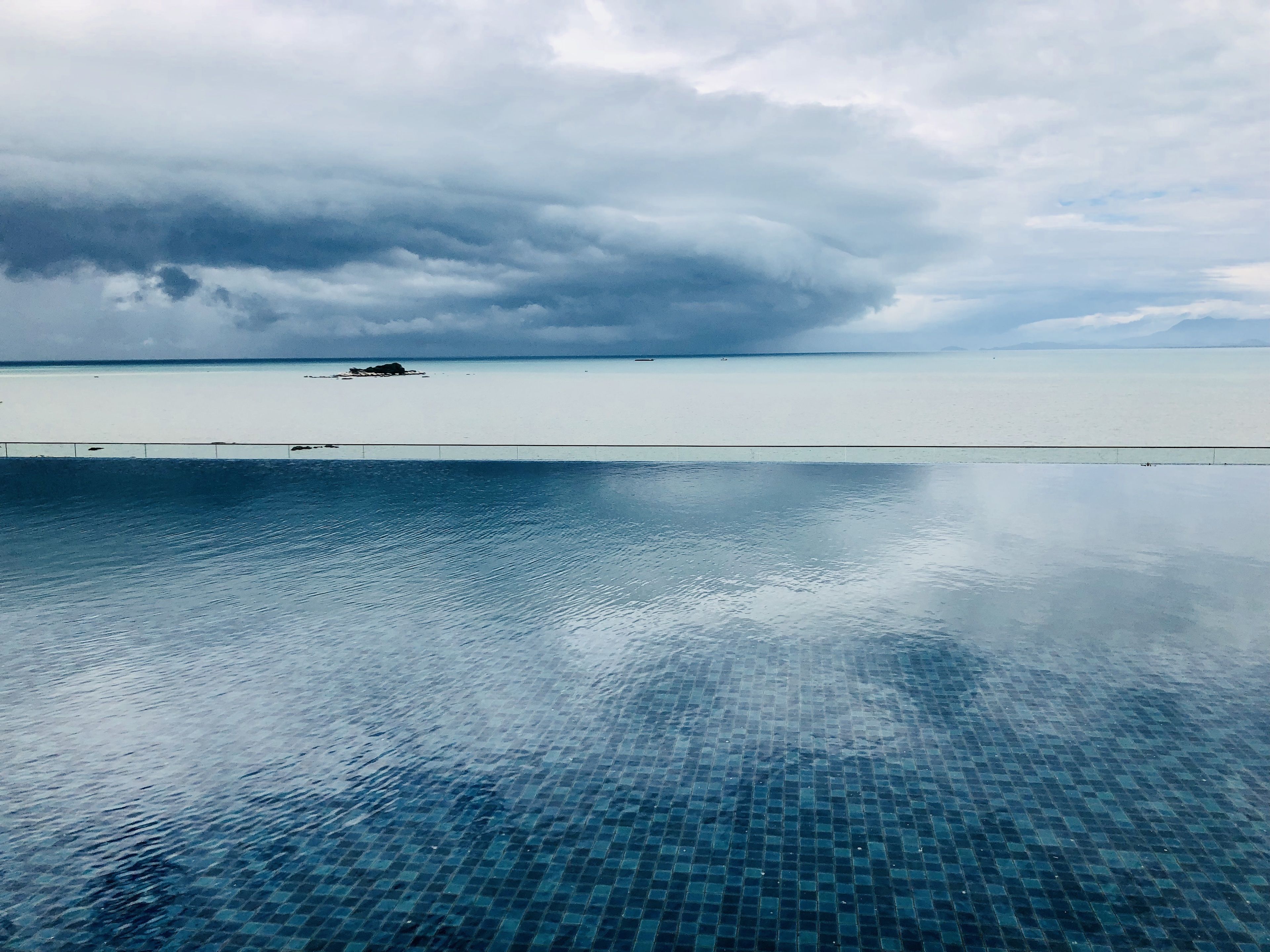 A clear view of a swimming pool with a tiled floor, a distant shoreline, and overcast skies.