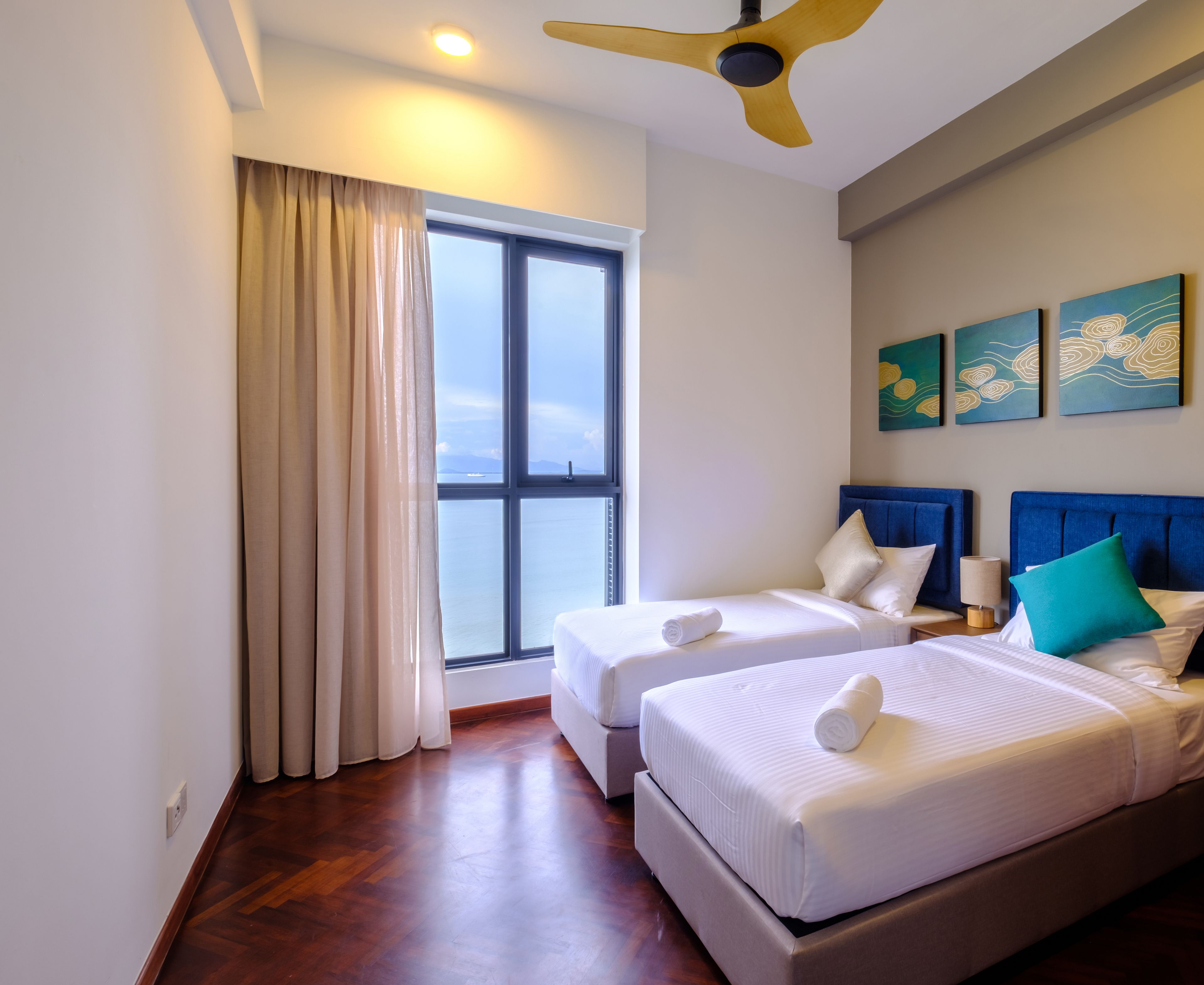A hotel room with two beds, a large window with curtains, a ceiling fan, and artwork on the wall.