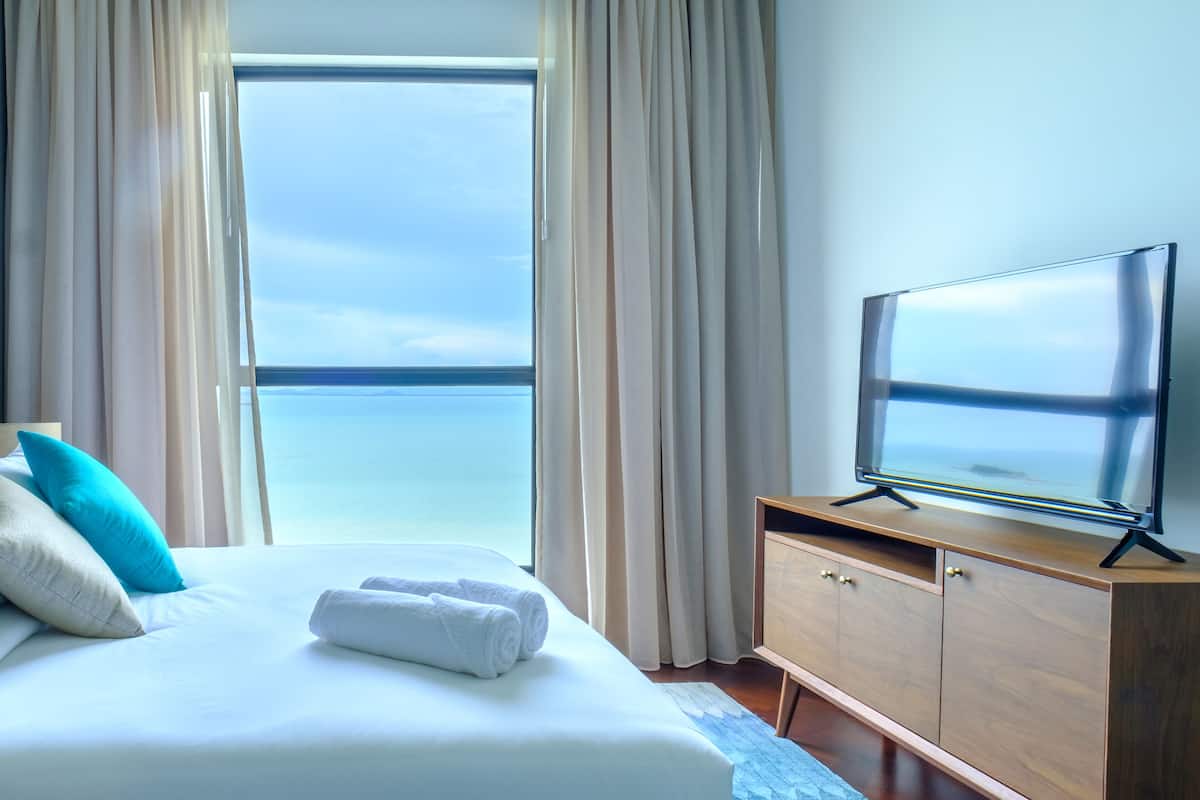 A hotel room with a bed, a TV on a wooden cabinet, and a view of the sea through large windows.
