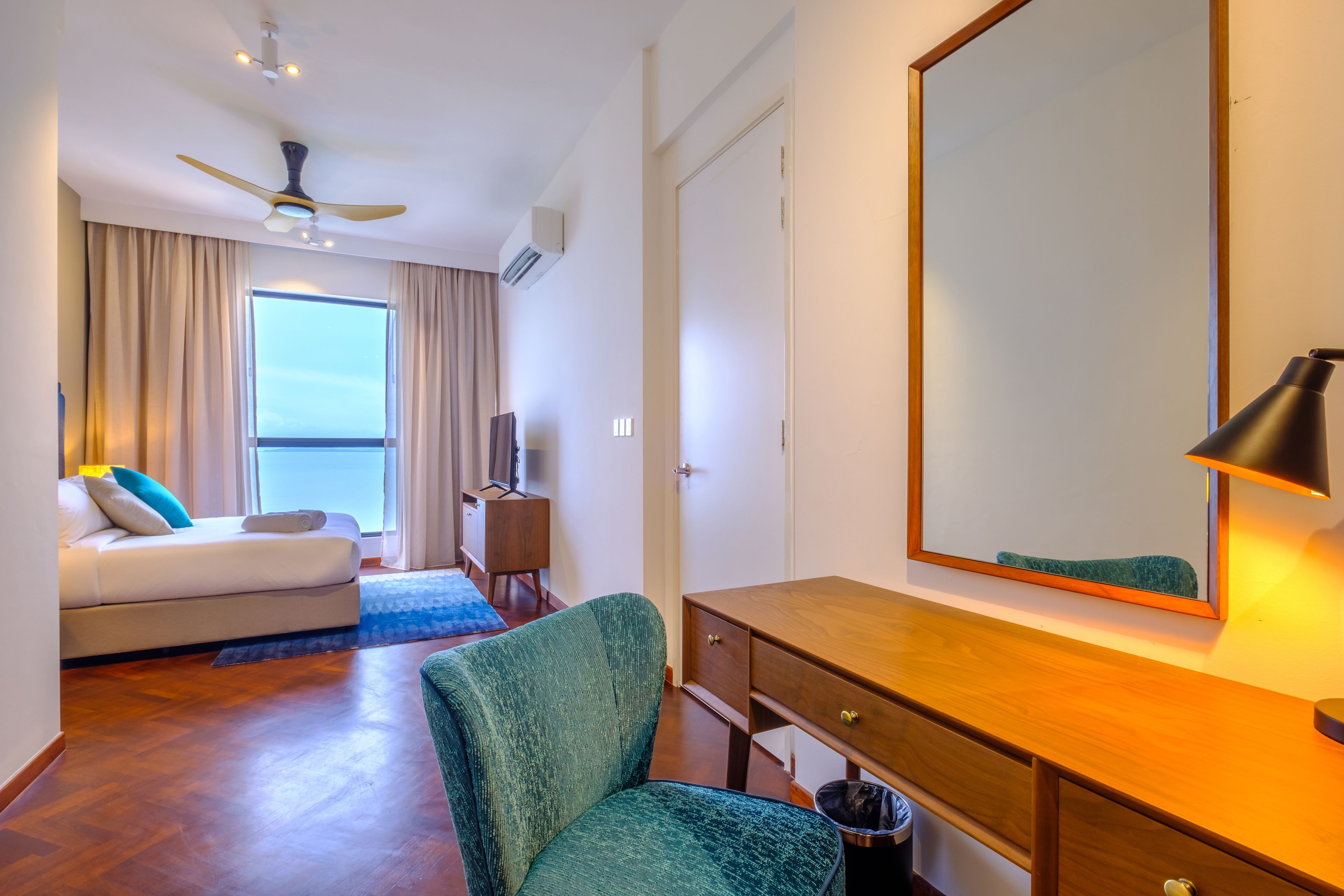 Une chambre d’hôtel comprenant un lit, un bureau, une chaise, un ventilateur de plafond, un grand miroir et une fenêtre avec des rideaux.
