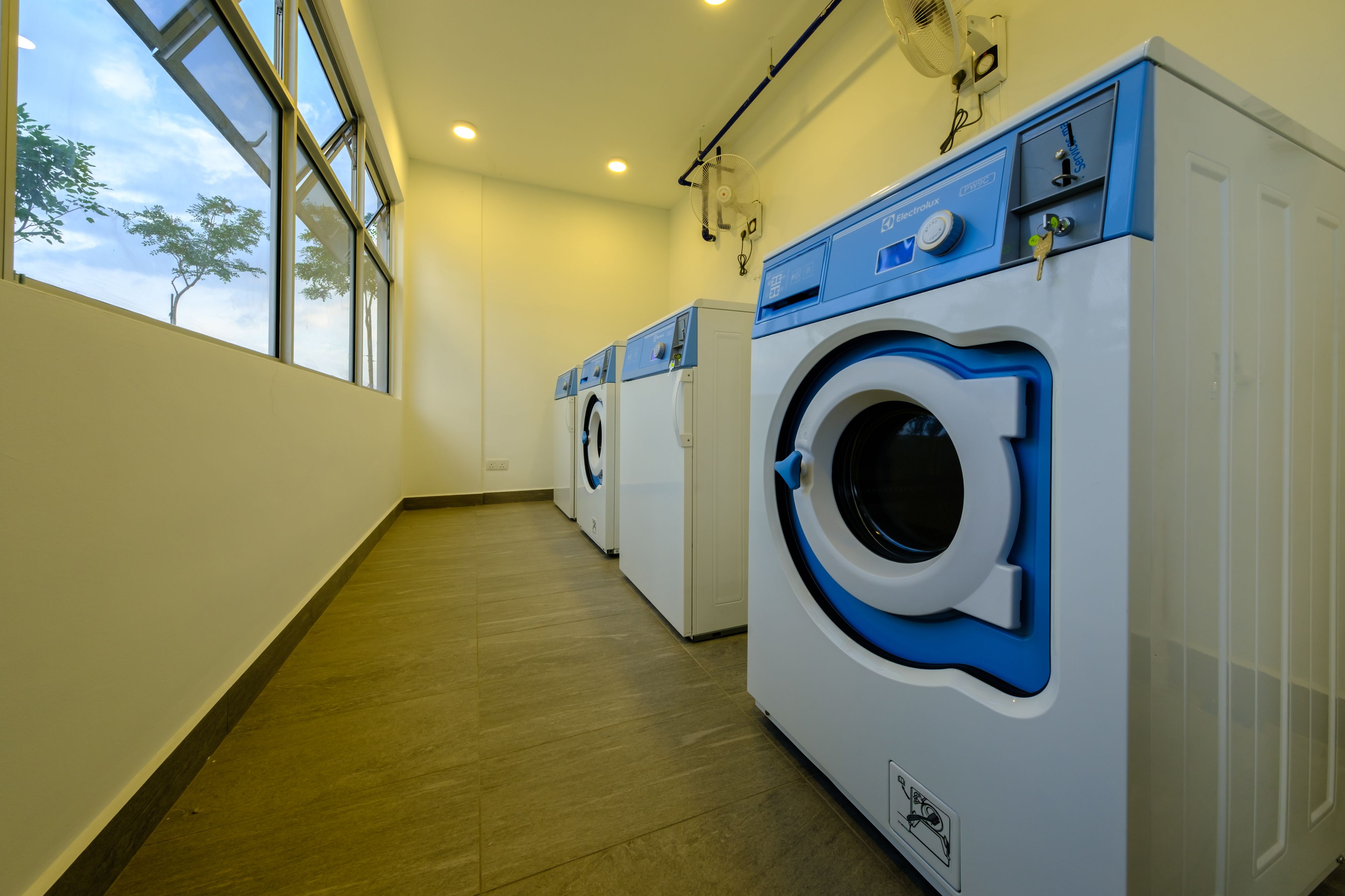 A laundry room with three front-loading washing machines, large windows, and a wooden floor.