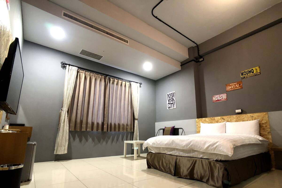 Une chambre d’hôtel moderne équipée d’un lit, d’une télévision, d’une fenêtre avec des rideaux et de décorations murales comprenant des noms de villes et une plaque d’immatriculation.