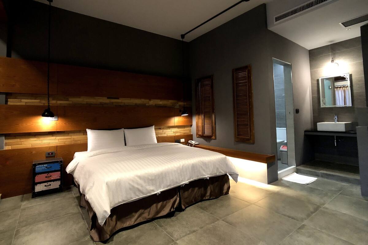 Une chambre d’hôtel moderne avec un grand lit, une tête de lit en bois, des lampes de chevet, une petite table de chevet, un miroir et une salle de bain équipée d’un lavabo et d’une douche.