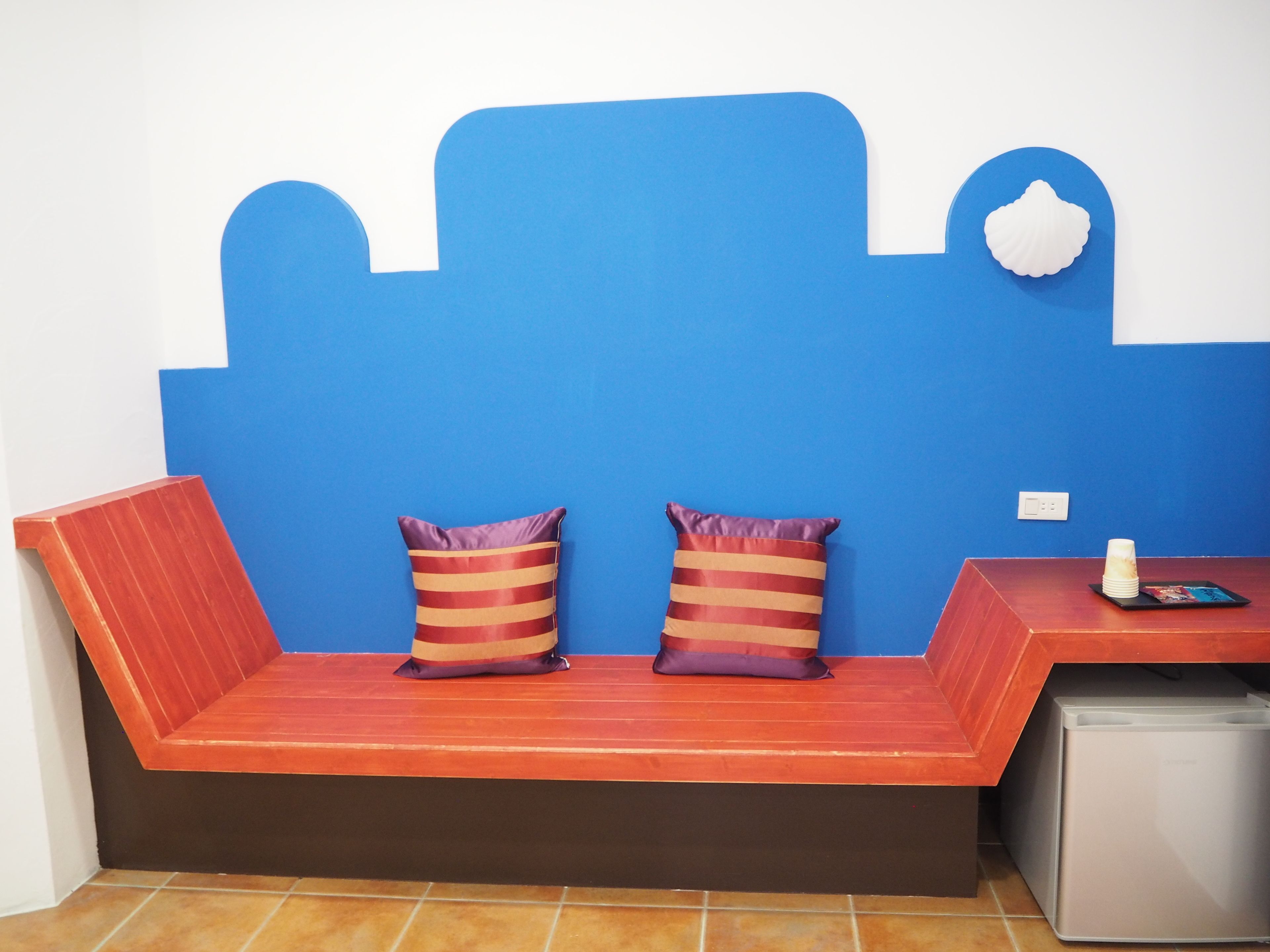 Una habitación con una pared azul, un banco de madera con dos almohadones a rayas, una mesita con una taza y un libro, y un refrigerador compacto.