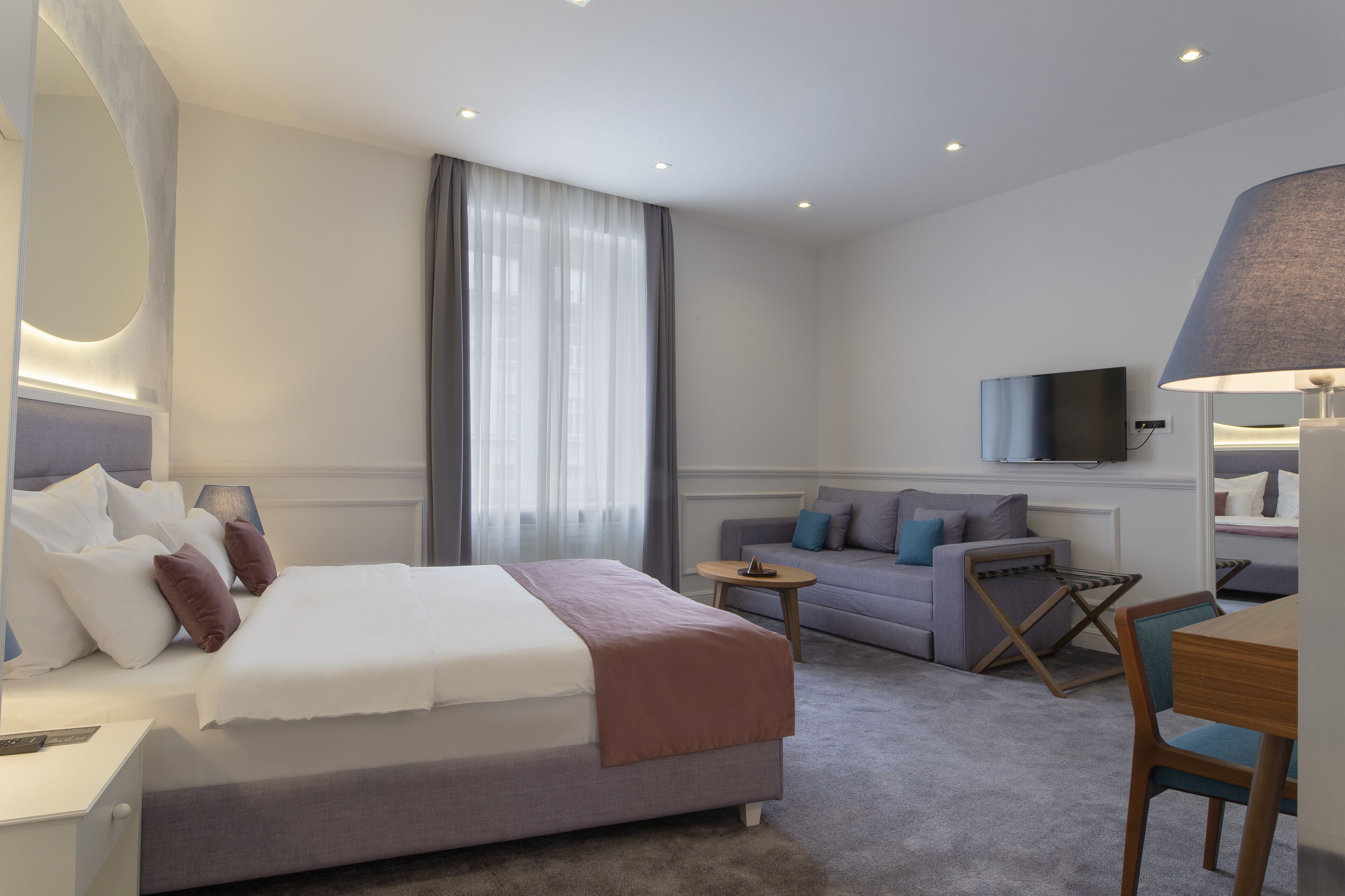 A modern hotel room with a large bed, a sofa, a TV, and a dining area.
