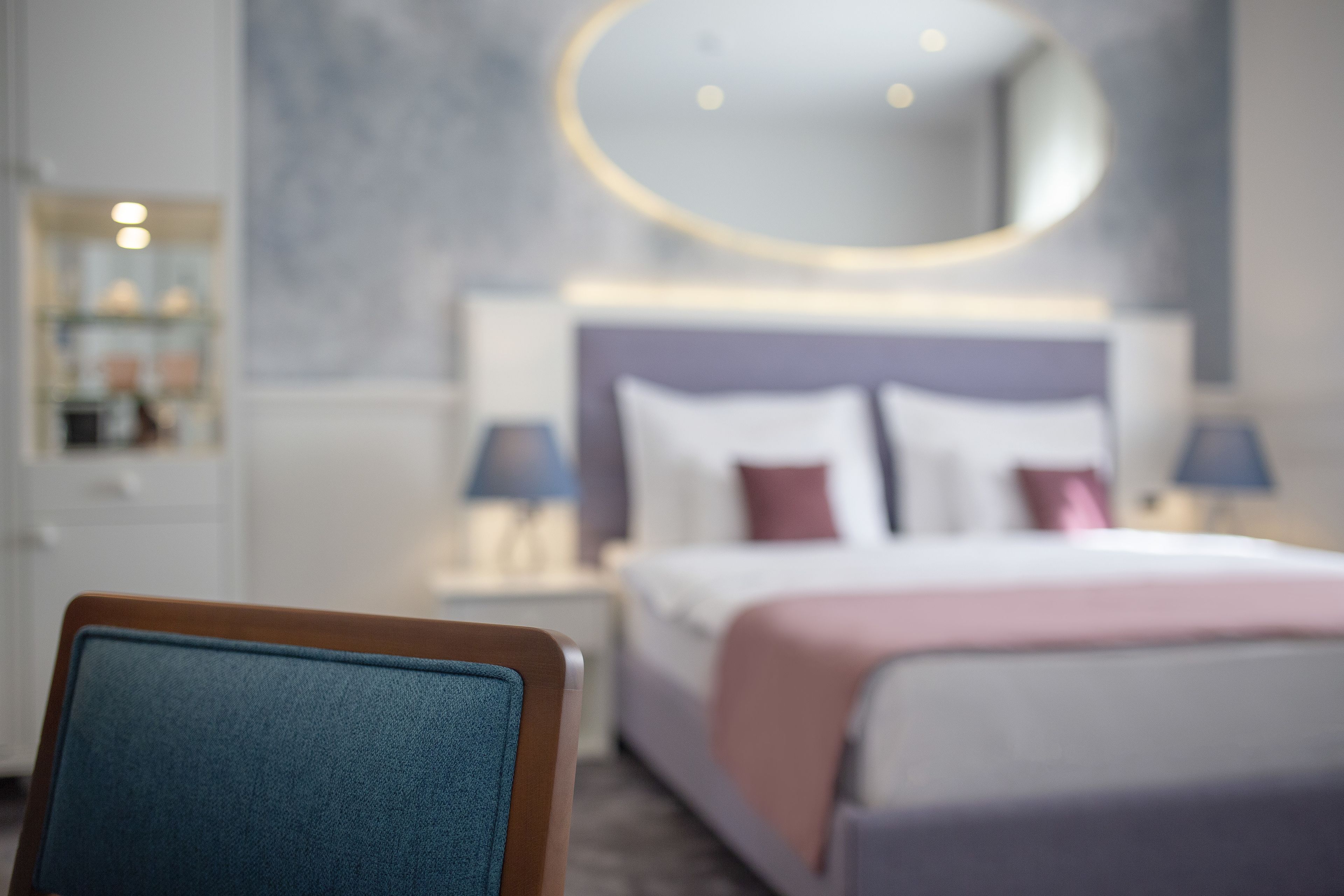 Una habitación de hotel moderna con una cama bien hecha, un espejo redondo sobre la cabecera y una mesita de noche con lámparas.