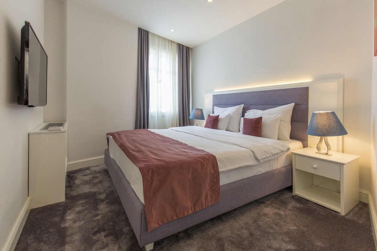 A hotel room with a bed, bedside tables, a TV, and a window with curtains.