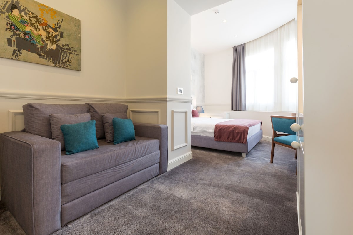 Una habitación de hotel con un sofá gris, una cama con una colcha roja, una silla azul y un cuadro en la pared.