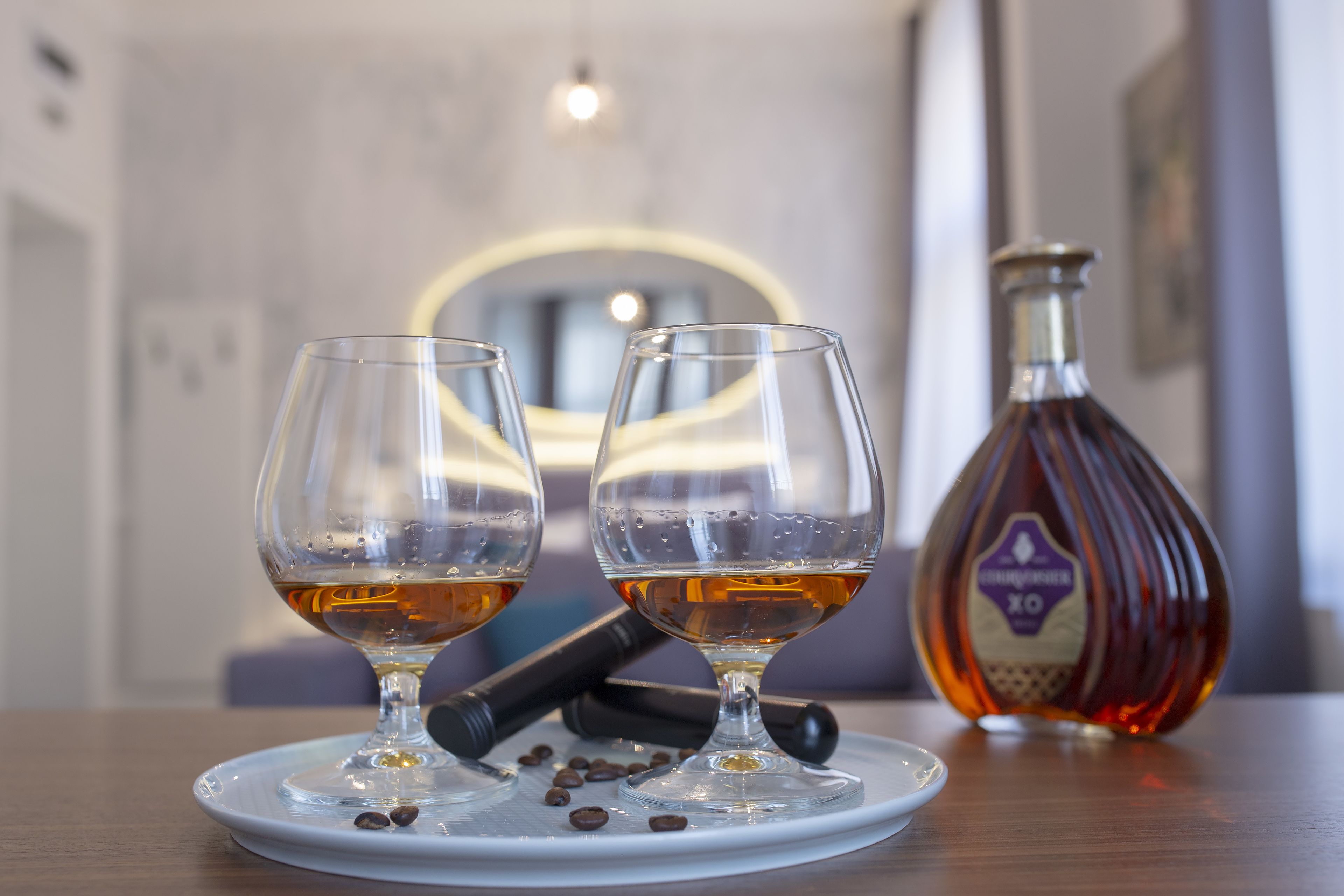 Dos vasos de líquido ámbar sobre un plato blanco, una botella de whisky XO y granos de café sobre una mesa de madera.