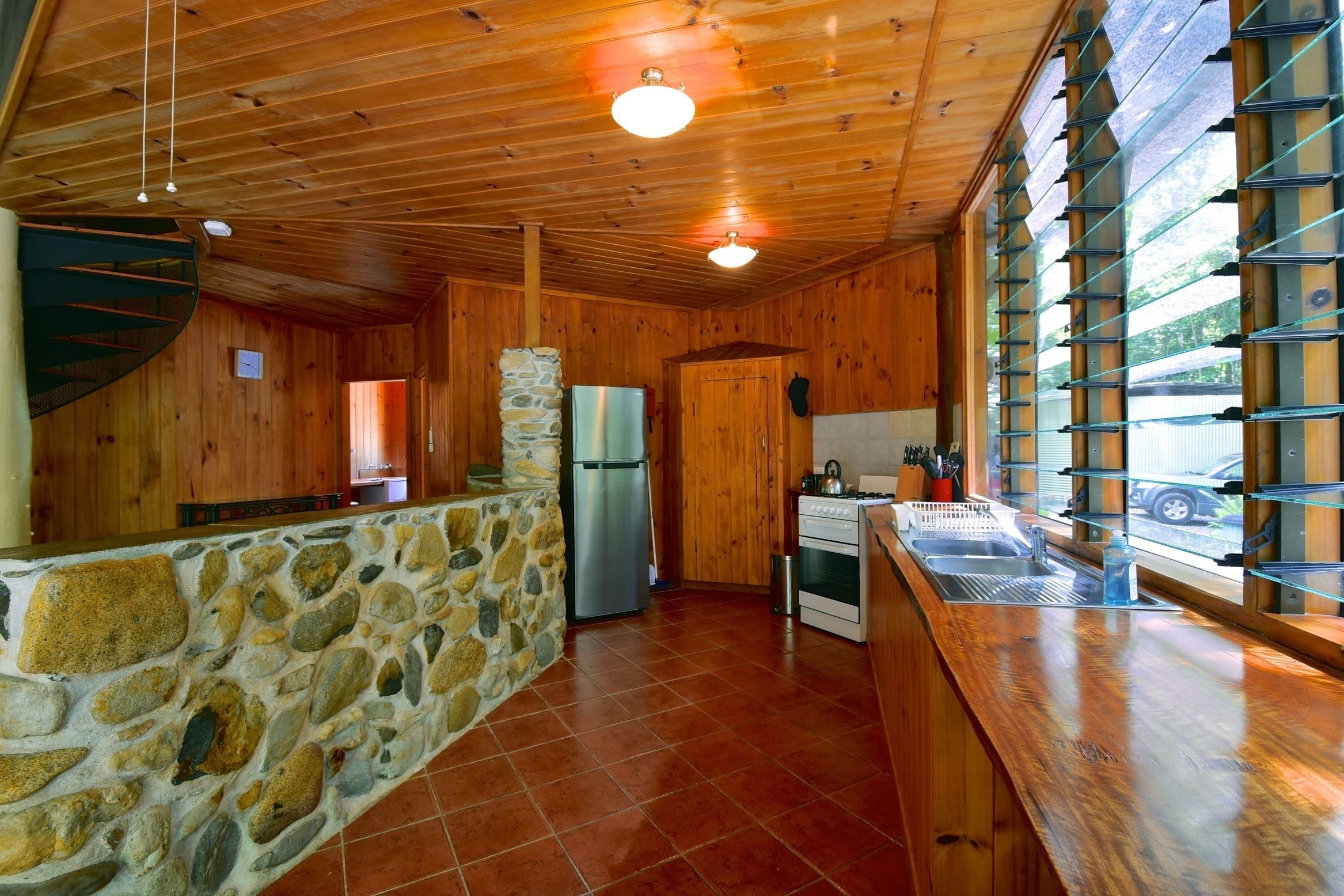Interior rústico de cabana com parede de pedra, teto de madeira e área de cozinha incluindo geladeira, fogão e pia.
