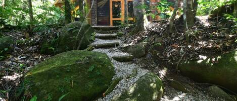 A wooden cabin with a stone staircase leading to a door, surrounded by a forested area with large moss-covered rocks.