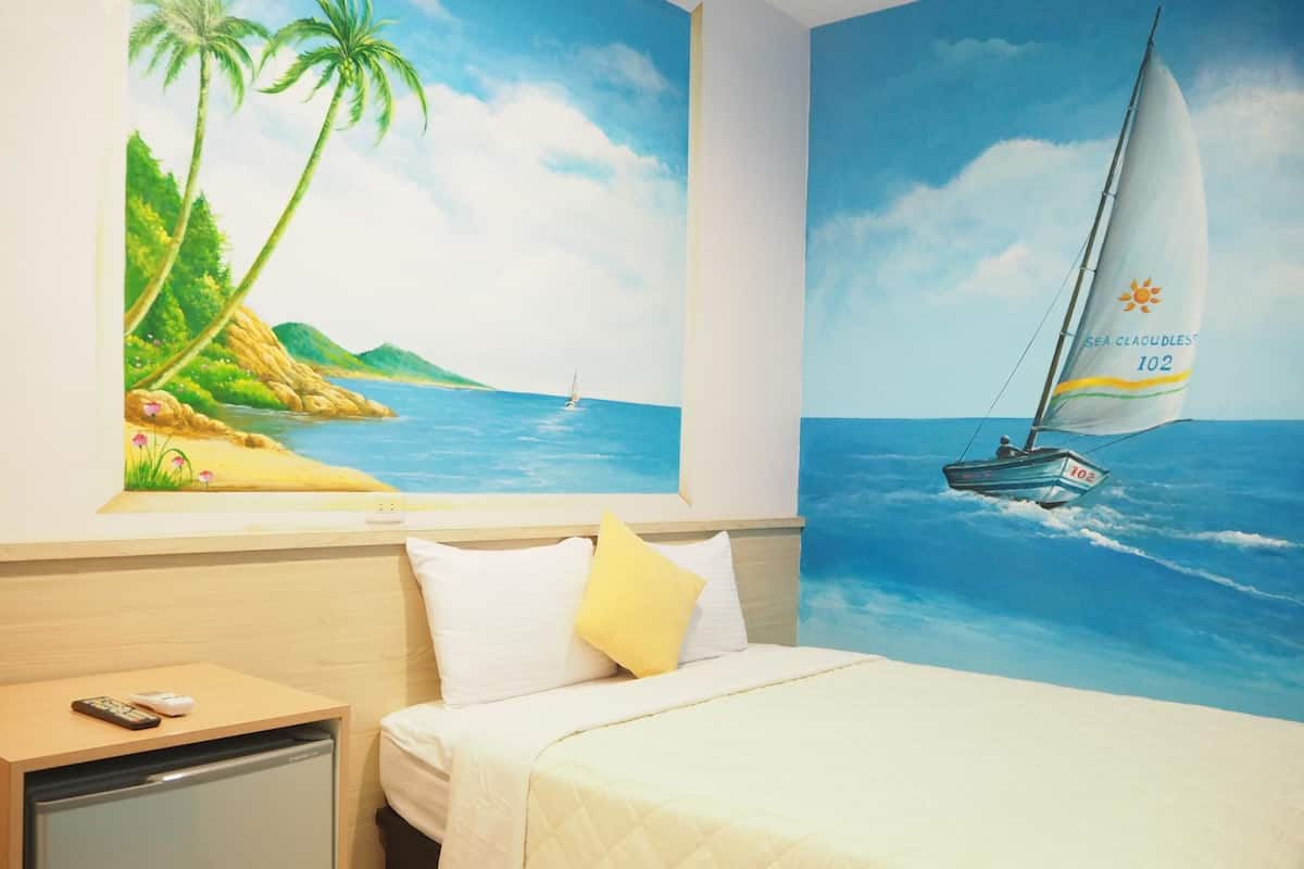 A hotel room with a bed, a nightstand with a remote control, and a wall mural depicting a beach scene with palm trees and a sailboat.