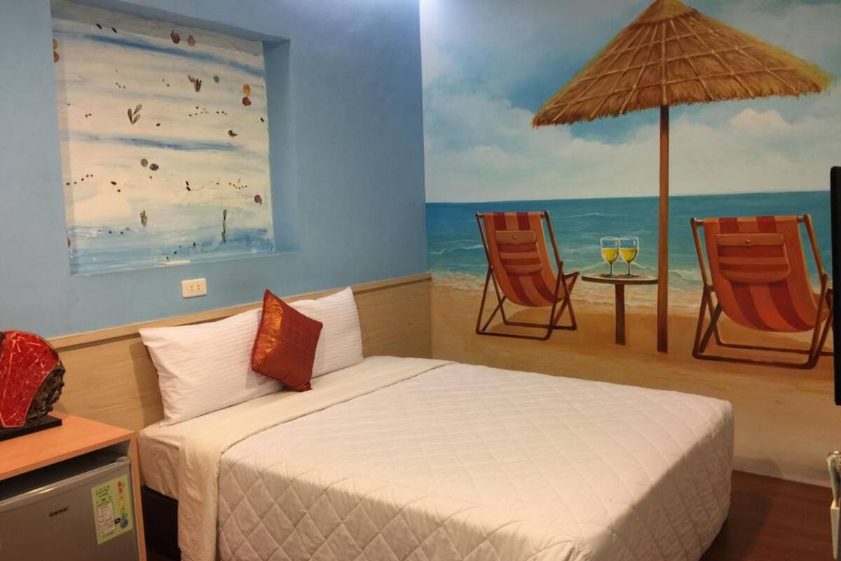 A hotel room with a bed, a small table with two glasses, and two beach chairs.