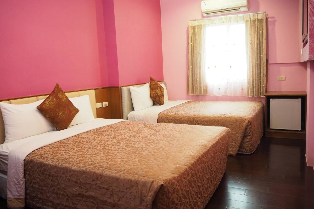 A hotel room with two beds, a small desk, an air conditioner, and a window with curtains.