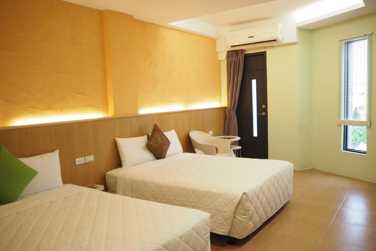 A hotel room with two beds, a small table, a chair, and an air conditioning unit.