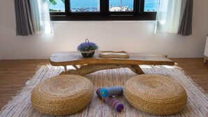 A wooden table with a potted plant and a decorative item, set on a woven rug with two round poufs, in a room with a view of the sea through large windows. - Najie (Donghe)