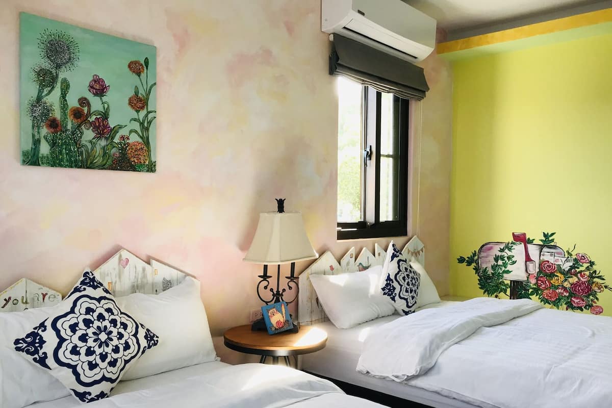 A bedroom with two beds, a window, an air conditioner, a lamp, and a painting on the wall.