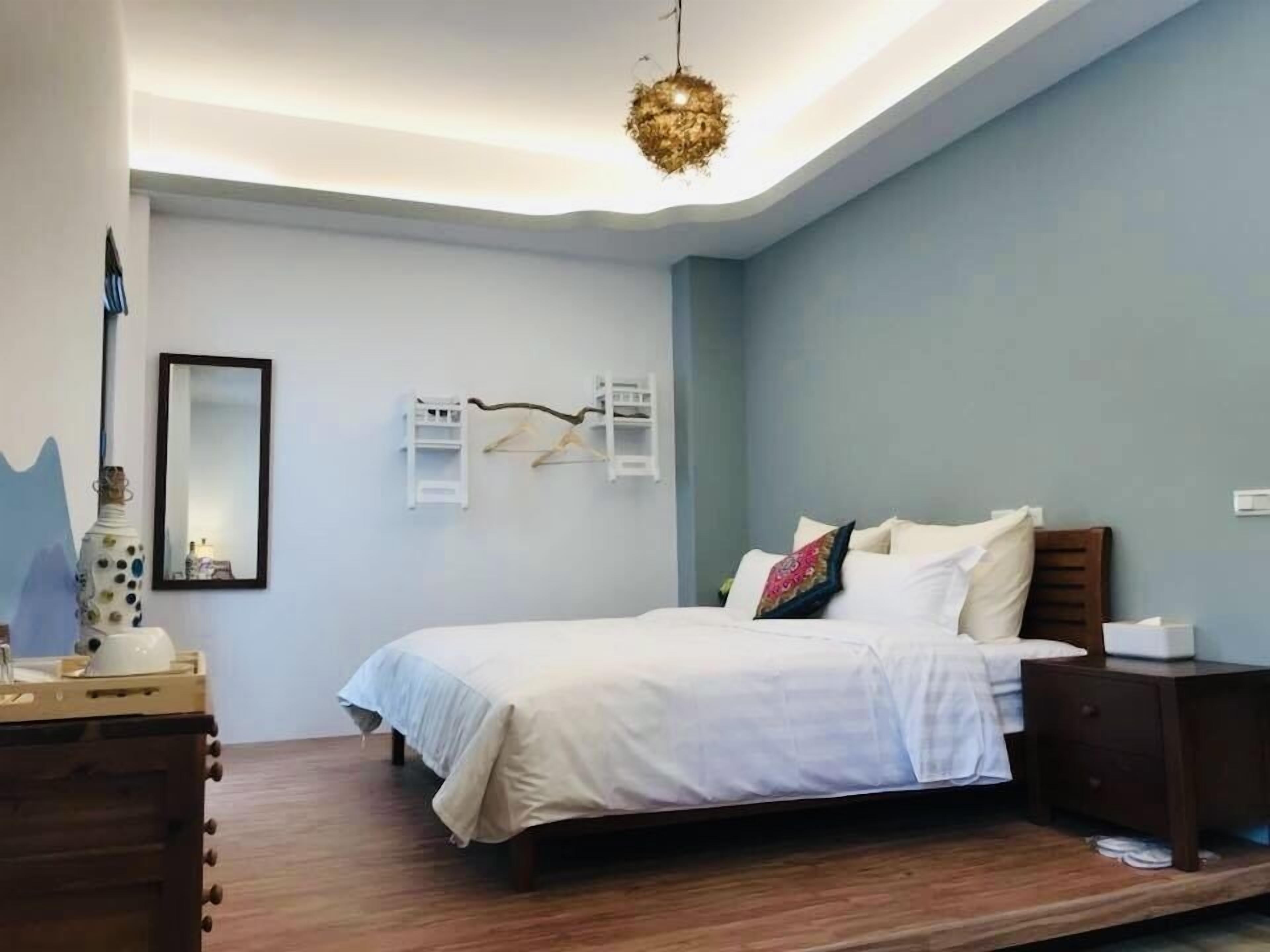 A bedroom with a bed, bedside tables, a hanging light fixture, a mirror, and a wall-mounted shelf.