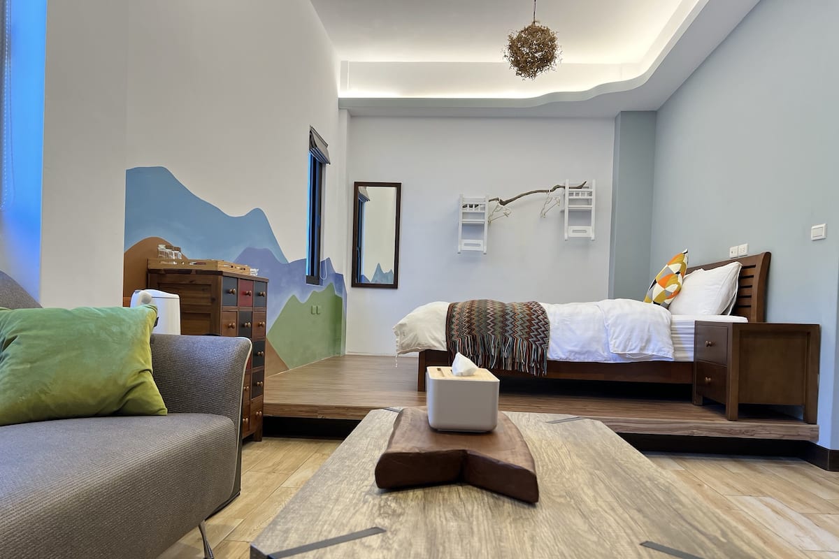 A modern living room with a sofa, a wooden coffee table, a bed with a wooden frame, and a wall with a mountain mural.