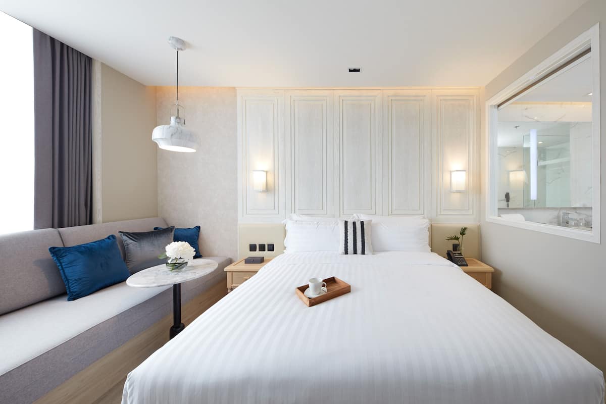 A hotel room with a large bed, a sofa with blue cushions, a small round table with a tray, and a hanging light fixture.
